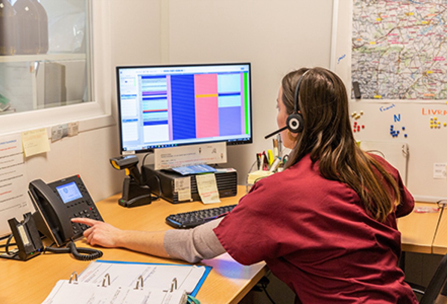 Une employée en blouse bordeaux, casque sur la tête, appuie sur une touche du téléphone de bureau devant un écran affichant un planning, près d'une carte murale.