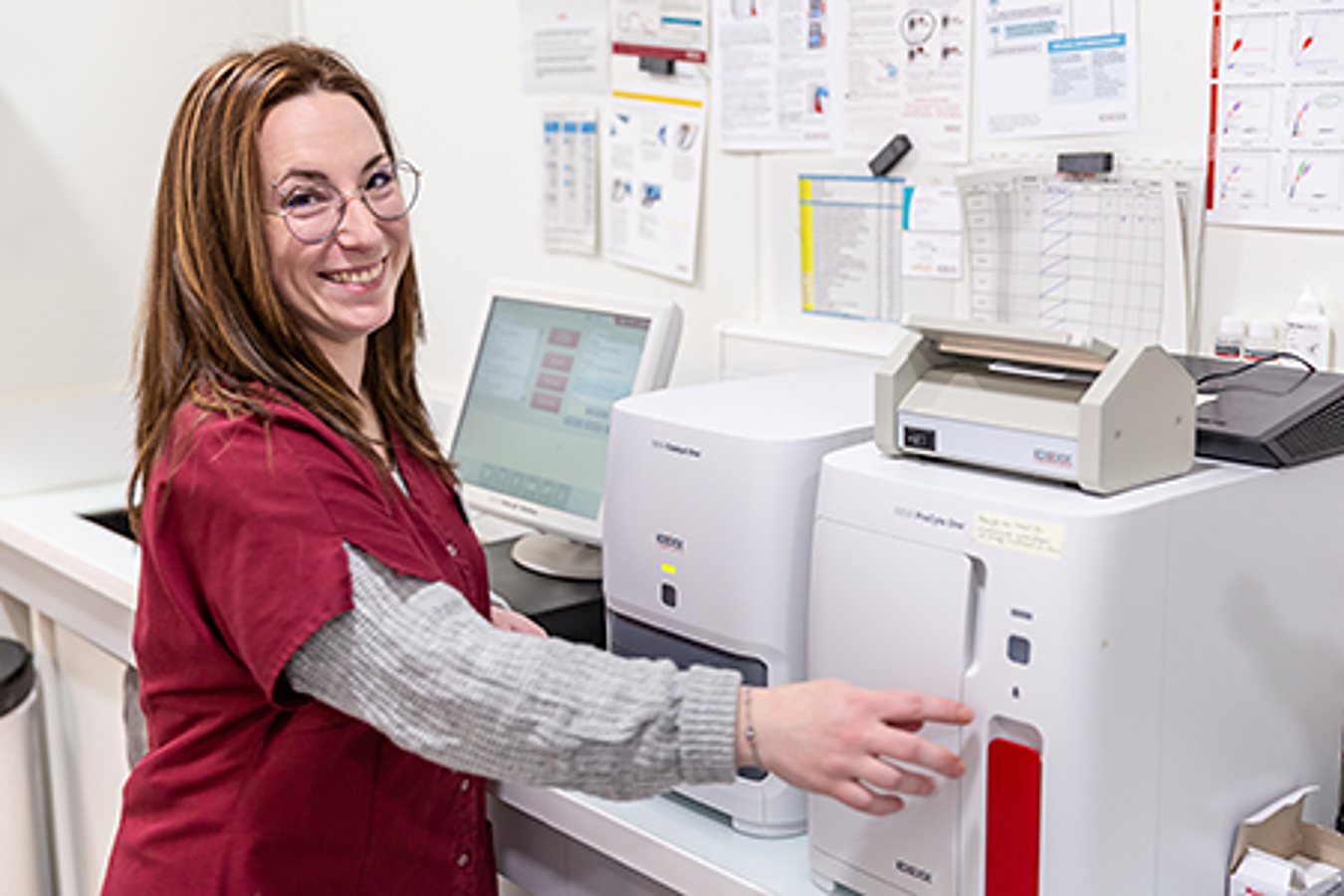 Une femme à lunettes en blouse bordeaux sourit tout en utilisant un appareil d'analyse posé sur un comptoir de laboratoire, avec un écran d'ordinateur derrière elle.