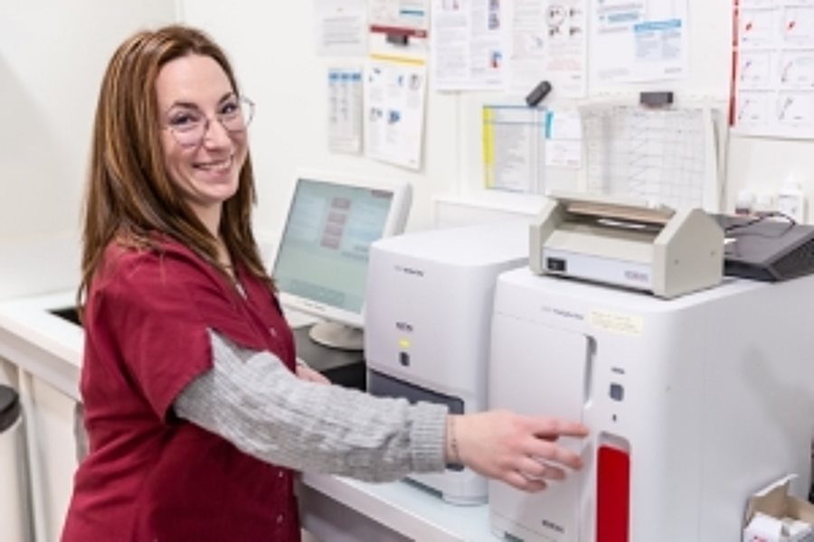 Une femme souriante portant des lunettes et une blouse bordeaux appuie sur un bouton d'un appareil d'analyse dans un laboratoire, avec un écran d'ordinateur et des documents affichés au mur derrière elle.