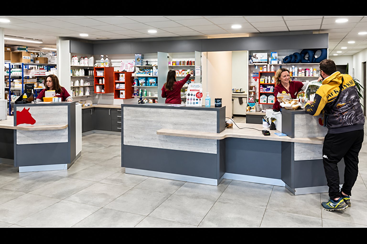 Intérieur moderne d'une pharmacie où des membres du personnel en tenue rouge servent un client debout devant les comptoirs gris et beiges.