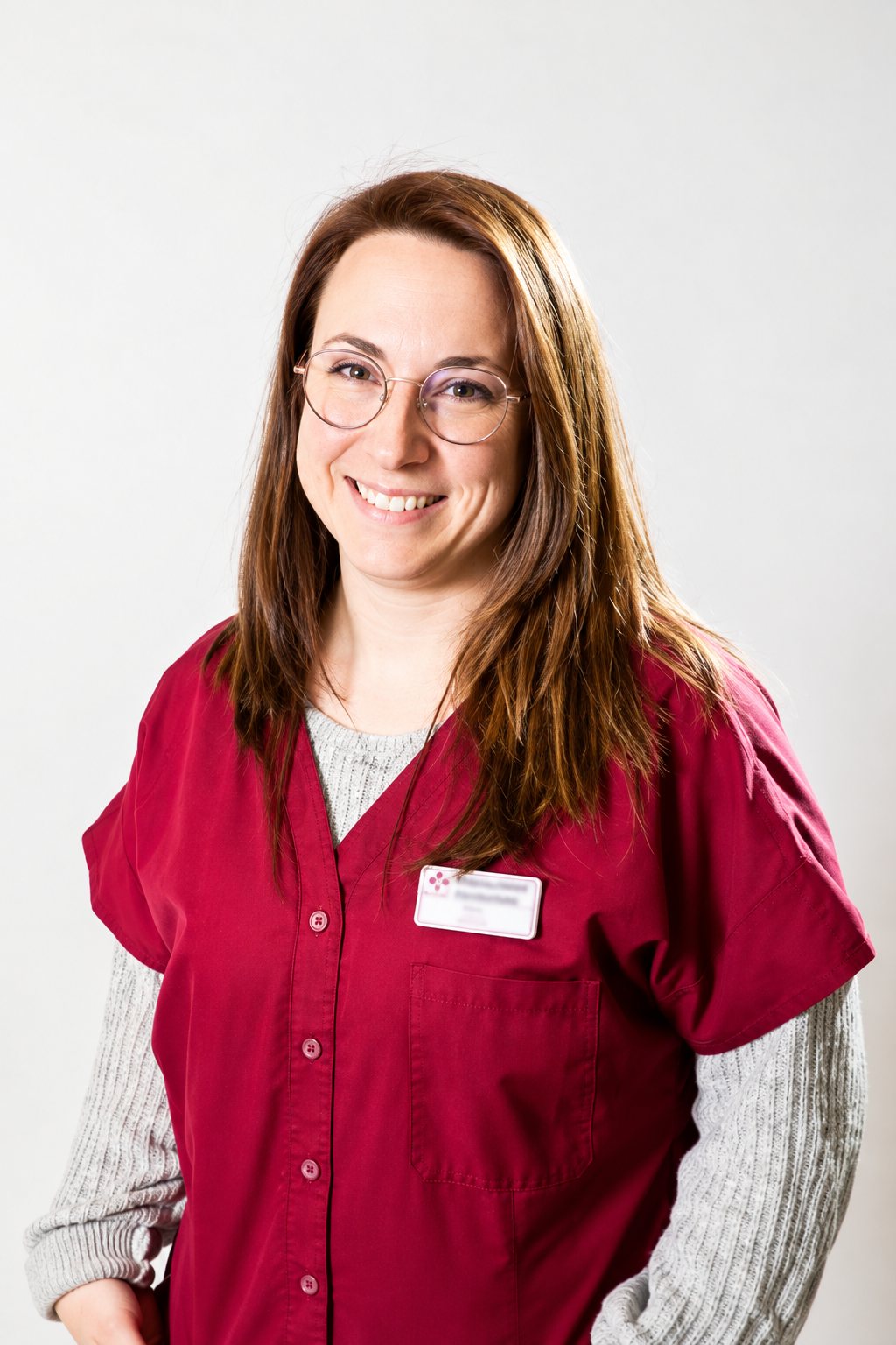 Une jeune femme souriante aux cheveux longs et aux lunettes rondes porte une blouse bordeaux avec un badge, devant un fond clair.