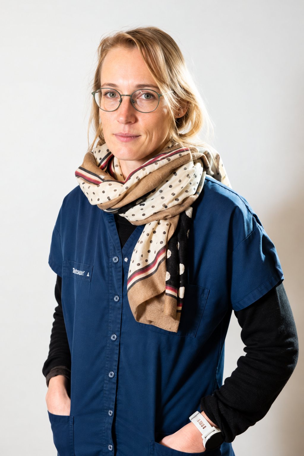 Une femme blonde à lunettes, vêtue d'une blouse bleu marine et d'un foulard à motifs beiges, pose devant un mur clair.