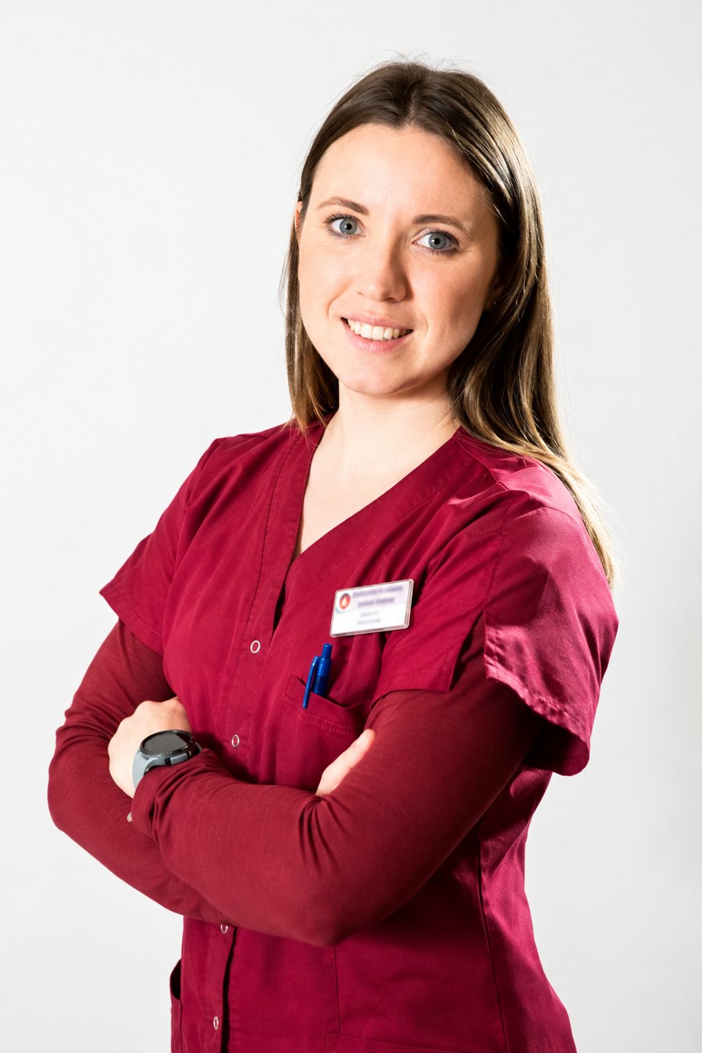 Une jeune femme aux longs cheveux châtains, vêtue d'une blouse bordeaux avec un badge, pose souriante les bras croisés devant un fond clair.