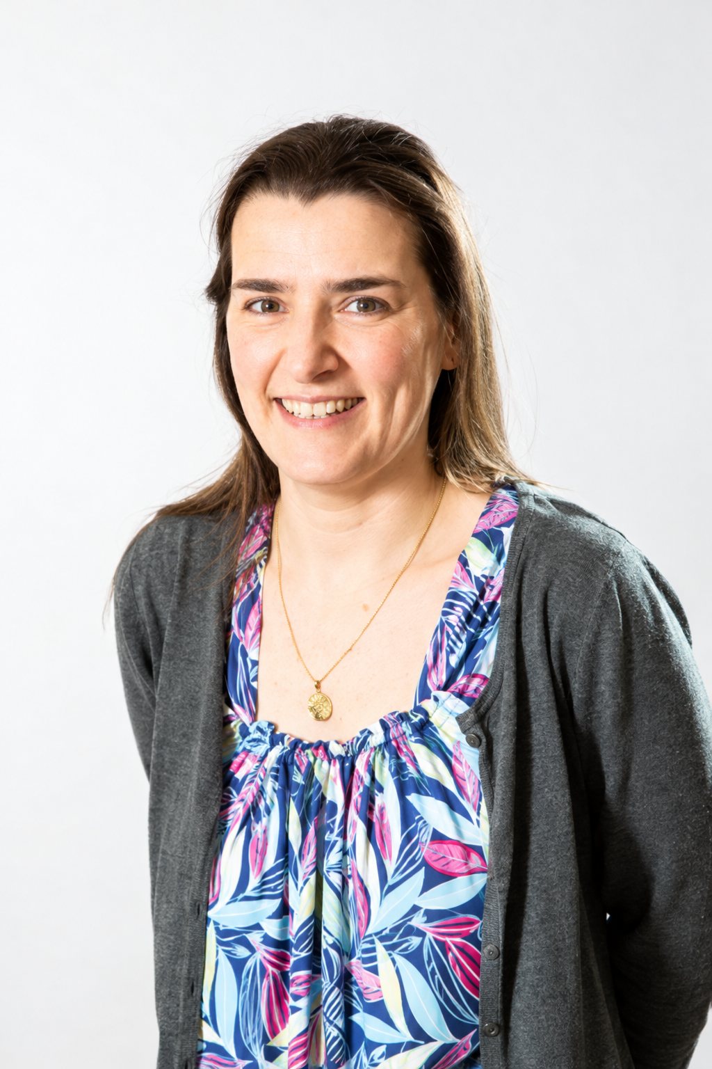 Une femme aux cheveux bruns mi-longs sourit devant un fond clair, vêtue d'un cardigan gris et d'un haut à motifs fleuris colorés.