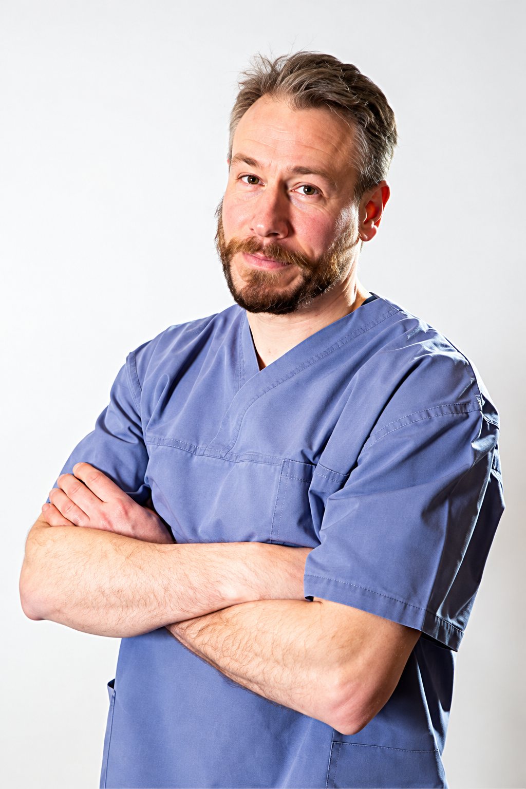 Un homme barbu en tenue médicale bleue pose les bras croisés devant un fond clair uni.