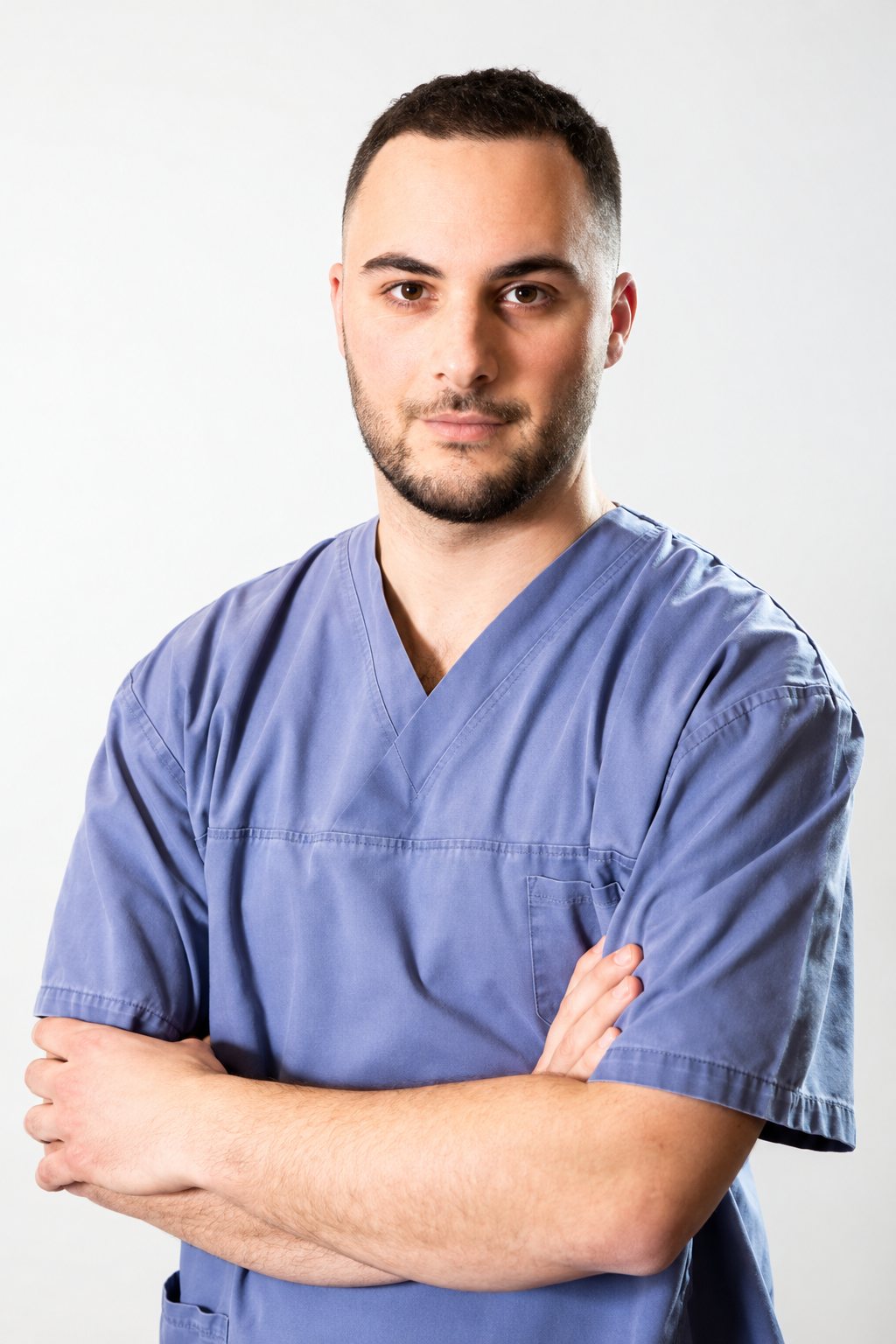 Un homme brun à la barbe courte, vêtu d'une blouse médicale bleue, pose les bras croisés devant un fond clair.