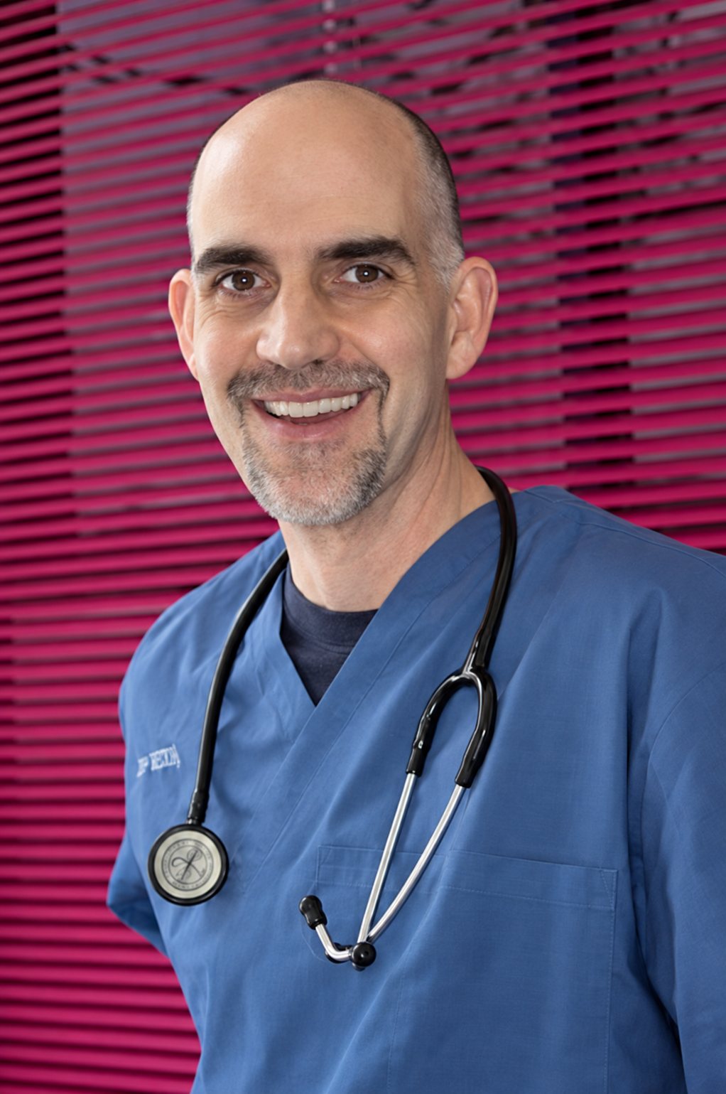 Un homme chauve et souriant, vêtu d'une blouse médicale bleue et portant un stéthoscope autour du cou, pose devant des stores rouges.
