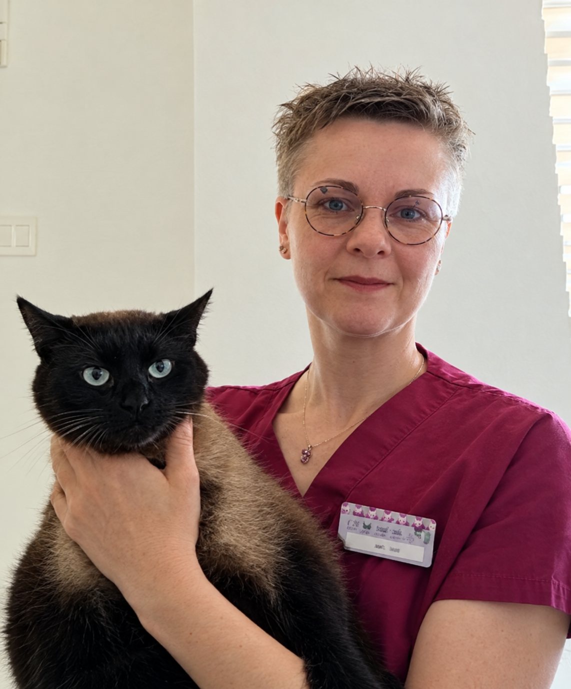 Une personne aux cheveux courts et à lunettes rondes, en blouse bordeaux avec un badge, tient dans ses bras un chat noir aux yeux verts.